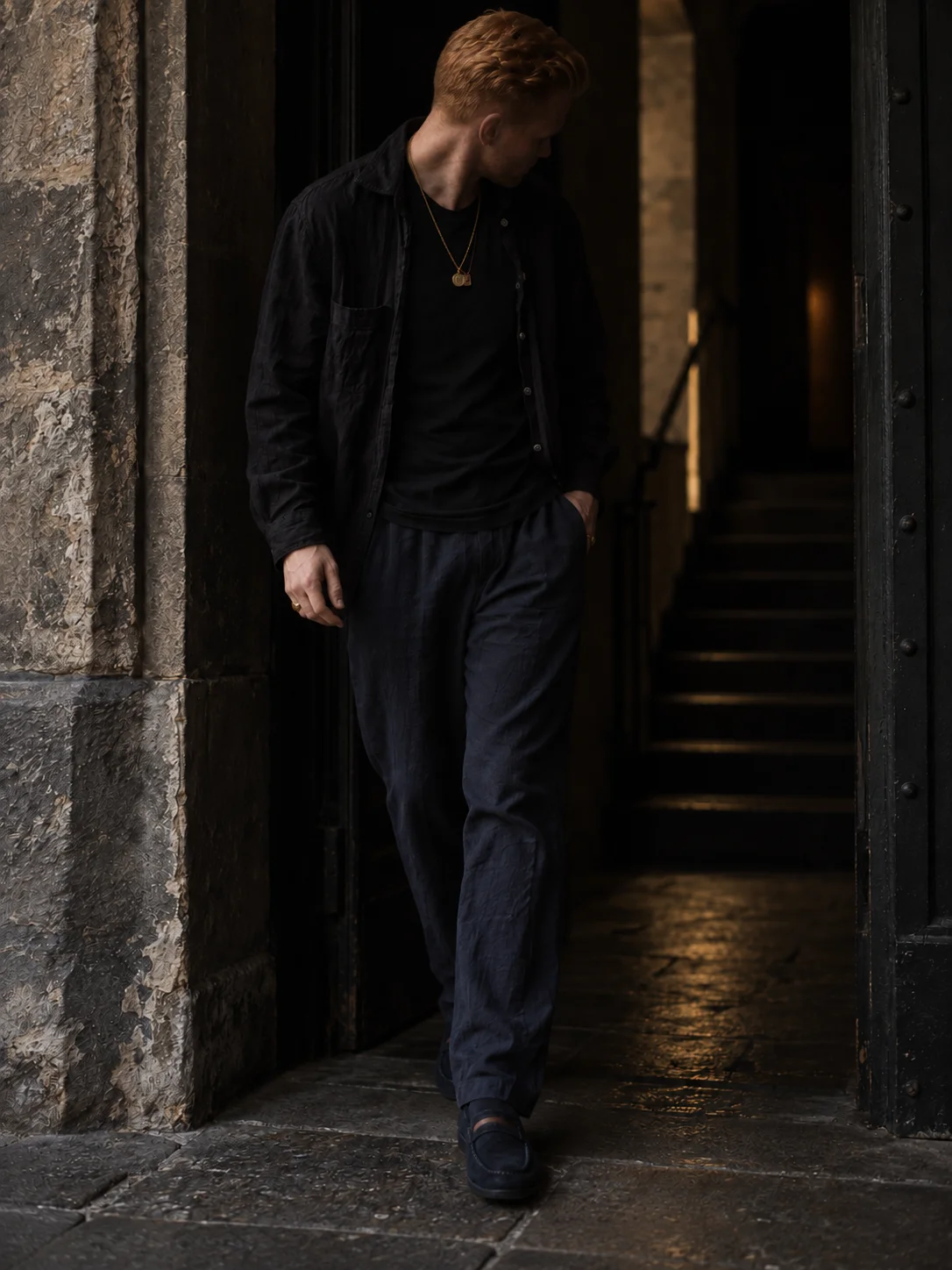 The same figure walking from a dark stone doorway in black linen, navy trousers and navy loafers