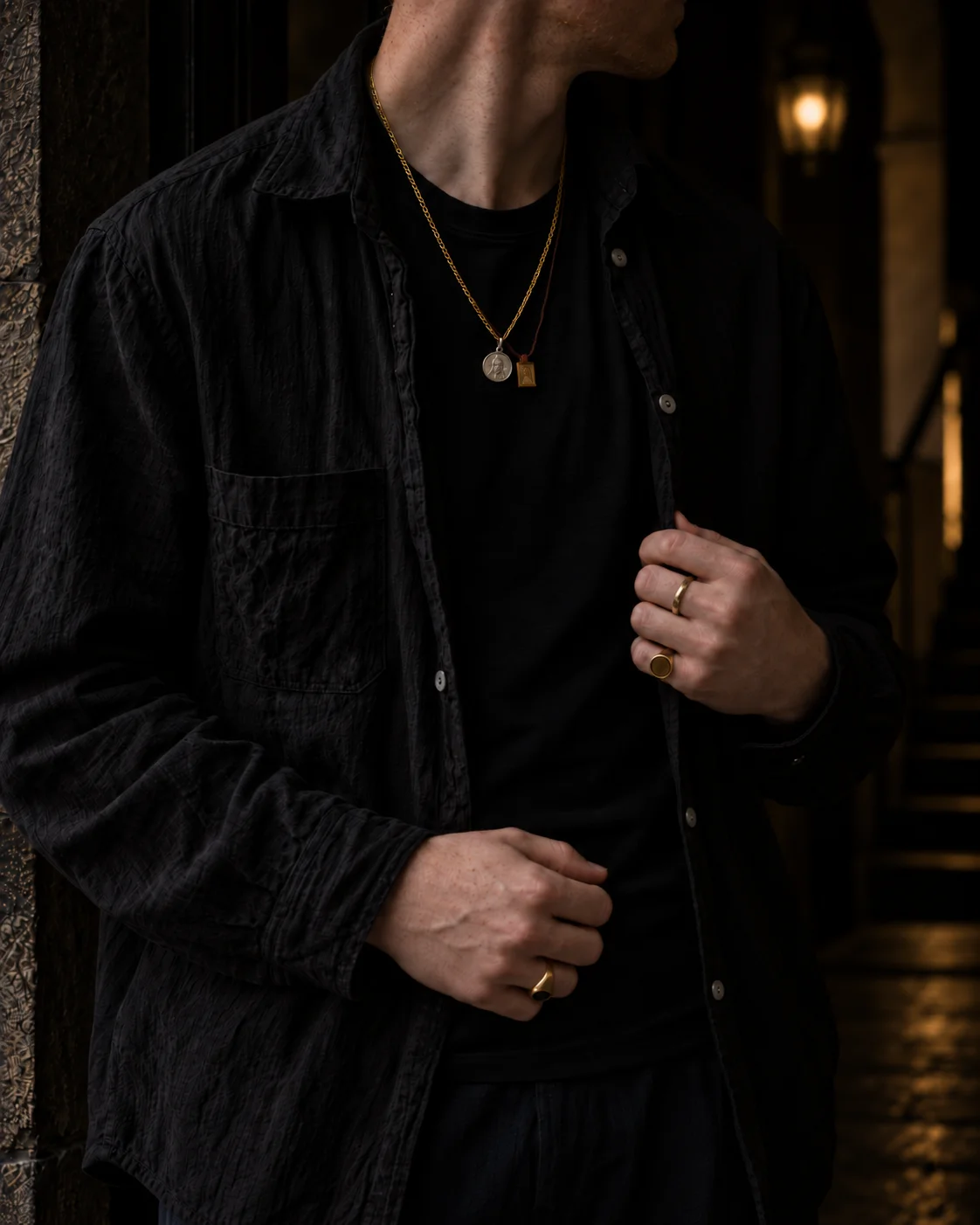 Close-up of a black linen overshirt, gold rope chain with coin and tablet pendants, and gold rings